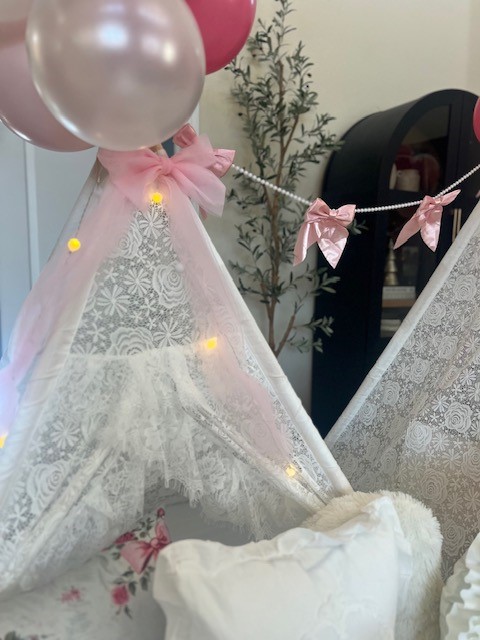 A close-up of the elegant pearls and bow garland draped across the floral-print teepees in a LoveShackFancy-inspired sleepover setup. The garland adds a touch of sophistication and vintage charm to the boho design.