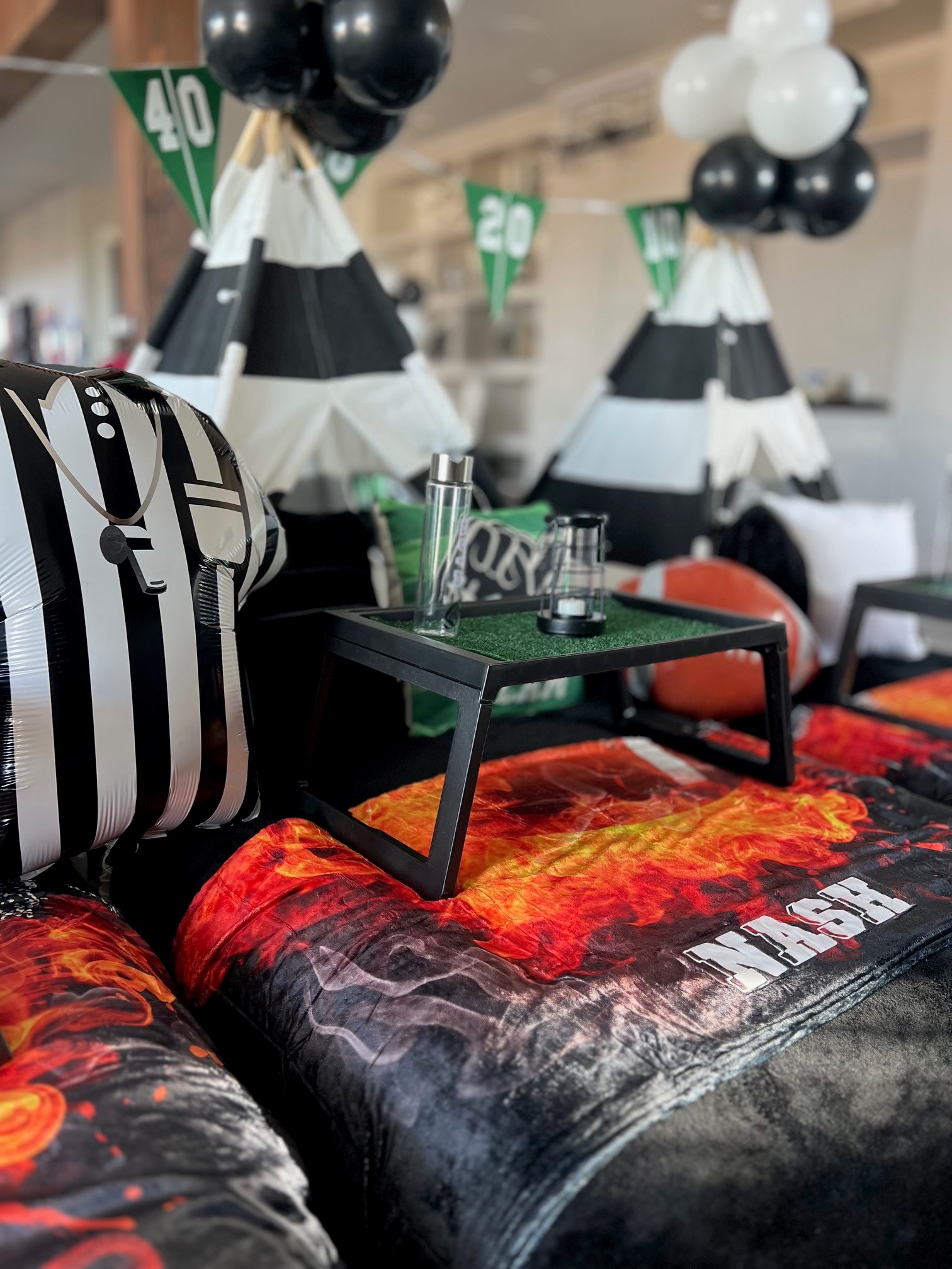 Close-up of a football-themed sleepover setup featuring custom football blankets, pillowcases, and mylar football balloons adorning stylish teepees.